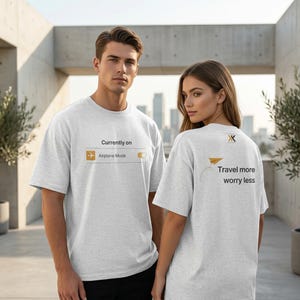 May include: Two people wearing light gray t-shirts. The front of the first shirt has the text "Currently on Airplane Mode" with a graphic. The second shirt has a paper airplane graphic and the text "Travel more worry less".