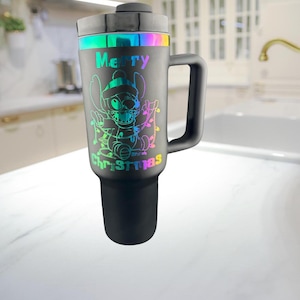 May include: A black stainless steel travel mug with a rainbow iridescent design featuring a cartoon character and the text "Merry Christmas".