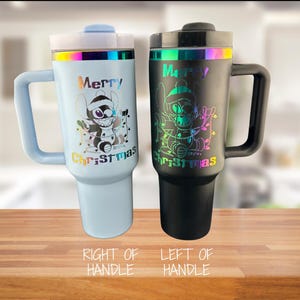 May include: Two stainless steel travel mugs with handles. The mug on the left is black with a rainbow iridescent finish and the mug on the right is light blue with a rainbow iridescent finish. Both mugs feature a cartoon character with the text "Merry Christmas".