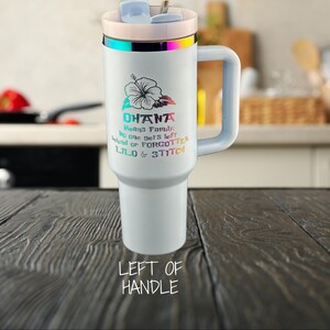 May include: White travel mug with a rainbow iridescent lid and handle. The mug has a floral design and the text "Ohana means family. No one gets left behind or forgotten. Lilo & Stitch".