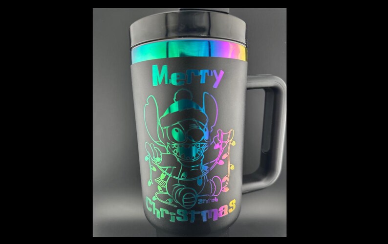 May include: Black travel mug with a rainbow iridescent band around the top. The mug has a handle and features a graphic of Stitch from Lilo & Stitch wearing a Santa hat and holding Christmas lights. The text "Merry Christmas" is printed on the mug.