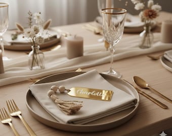 May include: A table setting with a beige linen napkin, gold cutlery, and a gold-rimmed glass. A gold name tag with the name "Charlotte" is on the napkin. The table is decorated with small vases of flowers and candles.
