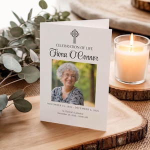 May include: A memorial card with the text "CELEBRATION OF LIFE" and "Fiona O'Connor" in elegant script. The card features a photo of a smiling woman and includes dates and location details. A lit candle sits nearby.
