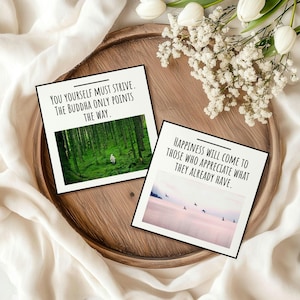 May include: Two square art prints with black borders on a wooden tray. One shows a forest scene with the text "YOU YOURSELF MUST STRIVE. THE BUDDHA ONLY POINTS THE WAY." The other shows a pink and blue landscape with the text "HAPPINESS WILL COME TO THOSE WHO APPRECIATE WHAT THEY ALREADY HAVE."