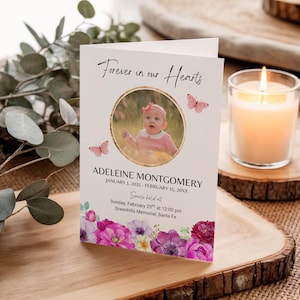 Op de afbeelding: Een herdenkingskaart met de tekst "Forever in our Hearts" en een foto van een baby. De kaart heeft een bloemenontwerp met roze en paarse bloemen. Een brandende kaars staat rechts van de kaart.