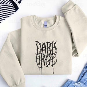 Könnte beinhalten: Cremefarbenes Sweatshirt mit dem Text "DARK URGE" in schwarzer, gotischer Schrift. Das Sweatshirt hat lange Ärmel und einen Rundhalsausschnitt. Der Text hat einen Tropfeffekt.