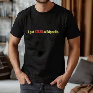 Op de afbeelding: Zwart T-shirt met de tekst "I got EDGED in Edgeville" in gele en rode letters. Het shirt heeft een ronde hals en lijkt van katoen te zijn. De persoon draagt het T-shirt.