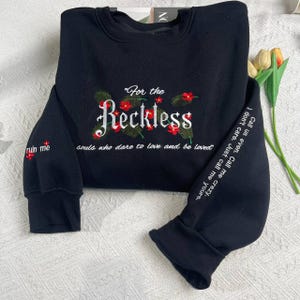 Puede incluir: Sudadera negra con texto bordado "For the Reckless souls who dare to love and be loved" y detalles florales. Las mangas tienen texto bordado adicional.
