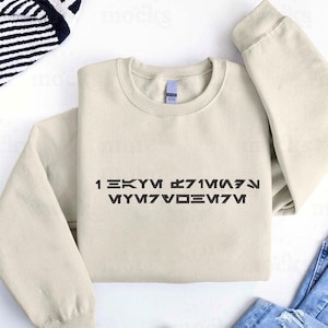 Può includere: Una felpa color crema con testo nero in un font stilizzato. Il testo recita "1 EKYM ℨ71M⋀⋋ MYM7V⌂⊾M7M". La felpa ha maniche lunghe e girocollo. L'indumento è piegato.