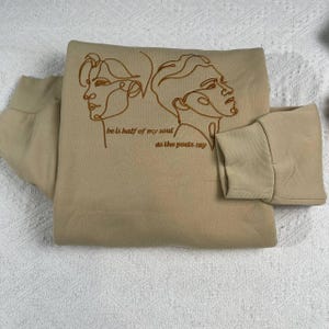 May include: A beige sweatshirt with a crew neck and long sleeves. The front features a line art illustration of two faces and the text "be is half of my soul / as the poets say" in a brown color.