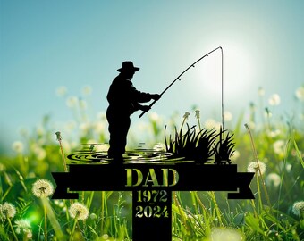 May include: Black metal garden stake featuring a silhouette of a person fishing, with the word "DAD" and the years "1972 2024". The background shows a sunny sky and a field of green grass and dandelions.