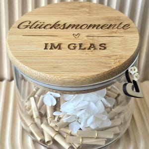 Könnte beinhalten: Ein durchsichtiges Glasgefäß mit einem runden Bambusdeckel, der mit dem Text "Glücksmomente IM GLAS" und einem kleinen Herzsymbol graviert ist. Im Inneren befinden sich zahlreiche kleine, gerollte beigefarbene Papierstreifen mit Goldringen und weiße Blütenblätter.