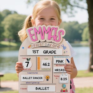 May include: Wooden sign with the name "EMMA'S" in pink letters, marking the first day of 1st grade. The sign includes information about the child's age, height (46.2 inches), favorite color (pink), and aspirations to be a ballet dancer.