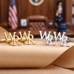 May include: Two pairs of cufflinks, one gold-toned and one silver-toned, each spelling out the letters "WRWR". The cufflinks are displayed on a tan ribbon, with a blurred background of a wooden desk and American flag.