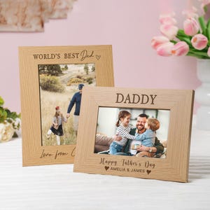 May include: Two light-coloured wooden picture frames. One frame reads "WORLD'S BEST Dad" and the other reads "DADDY". The frames contain family photos. The bottom of the "DADDY" frame reads "Happy Father's Day" and the names "AMELIA & JAMES".