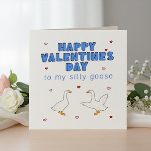 Può includere: Biglietto di San Valentino con la scritta "Happy Valentine's Day to my silly goose" in lettere blu. Il biglietto presenta due oche dei cartoni animati e piccoli cuori rosa. Il biglietto è su una superficie chiara con fiori e una candela sullo sfondo.