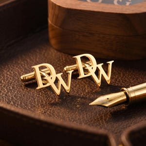 May include: Gold-colored cufflinks with the initials "DW" displayed on a brown leather tray. A wooden box and a gold fountain pen are also visible. The cufflinks are a classic accessory.