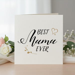 Peut inclure: Une carte de vœux crème avec les mots "BEST Muma EVER" en écriture noire et des accents dorés. La carte est décorée d'un cœur doré et d'éléments floraux. Une bougie allumée et des fleurs sont en arrière-plan.