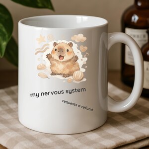 Può includere: Tazza in ceramica bianca con un'illustrazione di un criceto dei cartoni animati e la scritta "my nervous system requests a refund". Il criceto è marrone chiaro e sorridente. La tazza ha un manico curvo.