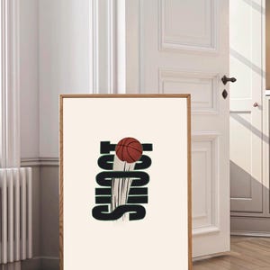 Puede incluir: Impresión enmarcada con una pelota de baloncesto y la palabra "Shoot". La pelota es marrón y el texto es verde oscuro sobre un fondo crema. El marco es de color marrón claro. La imagen está en una habitación con una puerta blanca.