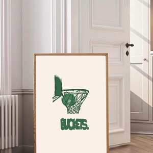 Puede incluir: Una impresión enmarcada que representa una pelota de baloncesto verde entrando en una red. La palabra "BUCKETS" está impresa en verde debajo de la red. La obra de arte está sobre un fondo crema, enmarcada en un marco de madera.