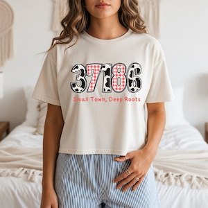 May include: Cream-colored t-shirt with the numbers "37186" in a mix of black and white cow print and red gingham patterns. The text "Small Town, Deep Roots" is printed below the numbers in pink.