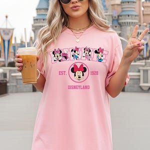May include: Pink t-shirt featuring Minnie Mouse designs and the text "EST. 1928 DISNEYLAND". The shirt has a crew neck and short sleeves. The design includes multiple Minnie Mouse illustrations.