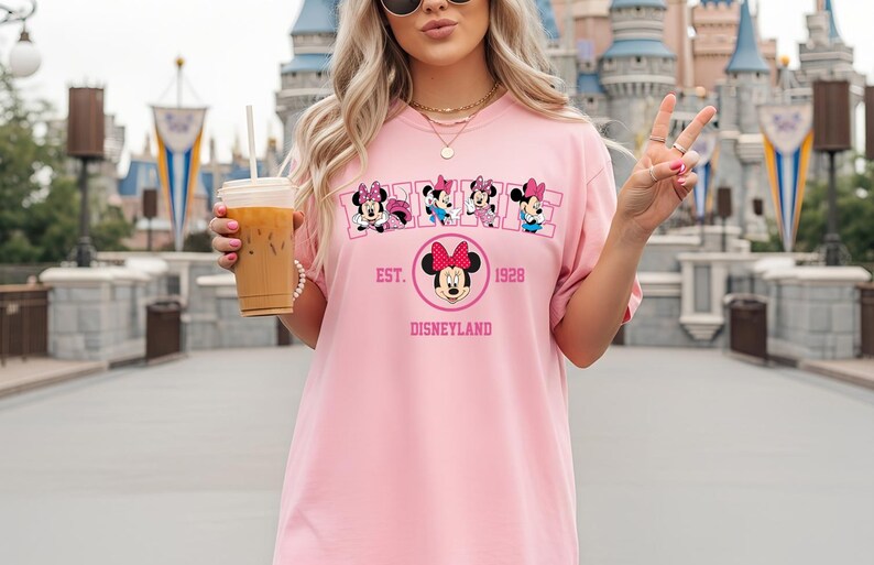 May include: Pink t-shirt featuring Minnie Mouse designs and the text "EST. 1928 DISNEYLAND". The shirt has a crew neck and short sleeves. The design includes multiple Minnie Mouse illustrations.