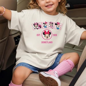 May include: A cream-colored t-shirt featuring a Minnie Mouse graphic with the text "EST. 1928 Disneyland". The shirt has a crew neck and short sleeves. The shirt is worn by a young child.