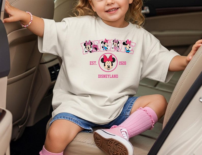 May include: A cream-colored t-shirt featuring a Minnie Mouse graphic with the text "EST. 1928 Disneyland". The shirt has a crew neck and short sleeves. The shirt is worn by a young child.