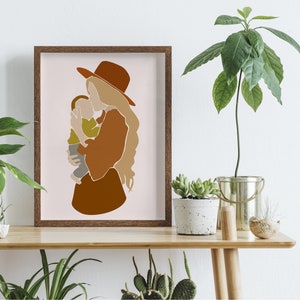 May include: A framed art print featuring an abstract illustration of a mother and child. The artwork uses warm earth tones, with a brown hat and dress, and a light green shirt for the child. The background is a soft pink.