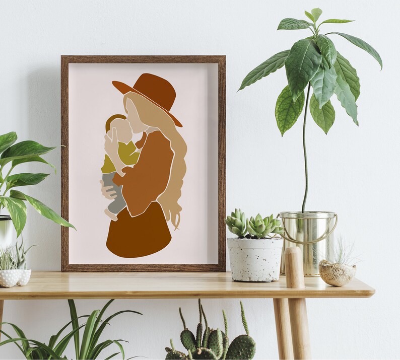 May include: A framed art print featuring an abstract illustration of a mother and child. The artwork uses warm earth tones, with a brown hat and dress, and a light green shirt for the child. The background is a soft pink.