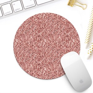 May include: A circular mouse mat with a rose gold glitter design. A white computer mouse sits beside the mat. A keyboard, gold binder clip, and pens are also in the image.