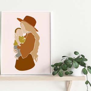 May include: Abstract art print featuring a mother and child in warm earth tones. The mother wears a brown hat and dress, holding the child in a tender embrace. The background is a soft pink, creating a minimalist and modern aesthetic. The artwork is framed in white.