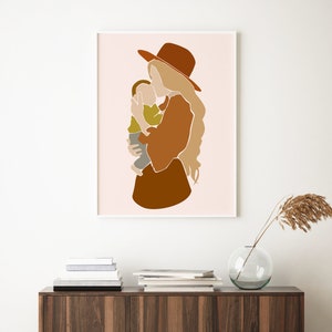 May include: Abstract art print featuring a mother and child in warm earth tones. The artwork depicts a woman in a brown hat and dress holding a child. The background is a soft pink, and the print is framed in white.