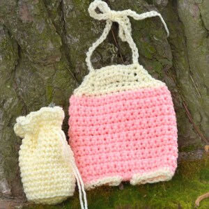 May include: A crocheted baby outfit in pink and cream with a matching crocheted bag. The outfit has a halter top style with a tie at the neck.