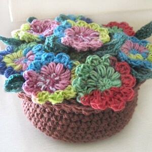 Puede incluir: Una maceta de flores de ganchillo con una base marrón y flores de ganchillo de colores en tonos de rosa, azul, verde y amarillo.