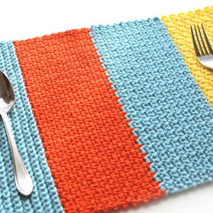 May include: A crocheted placemat with stripes of blue, orange, and yellow yarn. A silver spoon and fork are placed on the placemat.