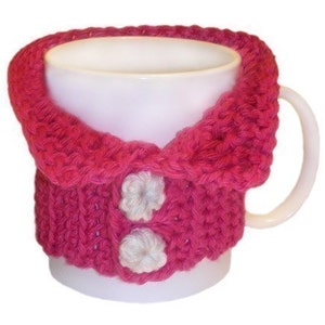 May include: A pink knitted mug cozy with white button details. The cozy has a collar-like design and is made with a soft, textured yarn.