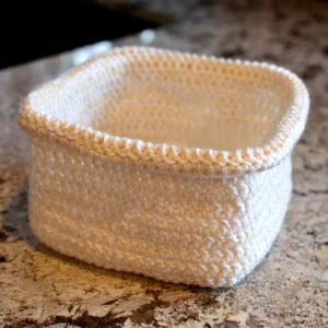 May include: A white crocheted basket with a square shape. The basket is made with a simple stitch pattern and has a smooth, even texture.