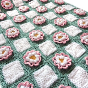 May include: A crocheted blanket with a checkered pattern in green and white. The blanket features pink flowers with white centers.