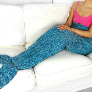 May include: A teal blue crocheted mermaid tail blanket with a white couch in the background.