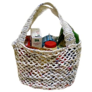 May include: A white crocheted mesh bag with a white handle. The bag is filled with groceries, including a box of strawberry fields cereal, a bottle of juice, and other items.