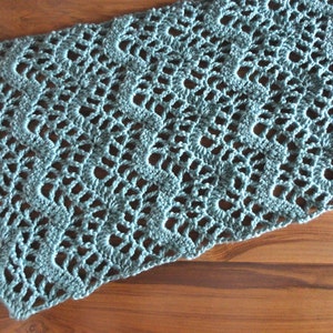 Puede incluir: Camino de mesa de ganchillo calado hecho a mano en color verde azulado apagado. El camino de mesa presenta un borde festoneado y un delicado patrón de encaje. El textil se muestra sobre una superficie de madera, mostrando su textura y diseño.