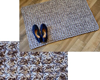 May include: A close-up of a crocheted rug with a brown and white color scheme. The rug is made with a textured stitch that creates a raised pattern. The rug is lying on a wooden floor.