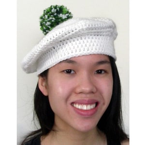 May include: A white crocheted beret with a green pom-pom on top. The beret is worn by a person with dark hair and a smiling face.