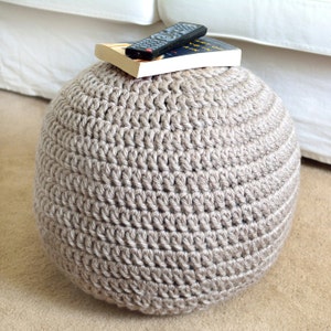 May include: A large, round, beige crocheted ottoman with a book and a remote control on top. The book is titled "Art of Living in a  Pain"