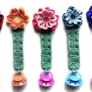 May include: Six crocheted flower pacifier clips with different colored flowers and matching pacifier clips. The clips are made of green yarn and the flowers are made of different colors of yarn. The pacifier clips are attached to the flowers with a metal ring.