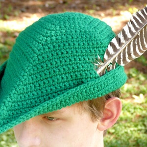 May include: A green crocheted hat with a pointed brim and a feather accent. The hat is worn by a person with brown hair.