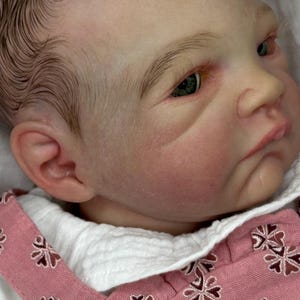 Może przedstawiać: Zbliżenie na lalkę niemowlęcą o realistycznych rysach twarzy, w tym malowanych włosach, zielonych oczach i różowej cerze. Lalka jest ubrana w różowo-biały strój z kwiatowym wzorem.